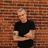 a photo of Henry Rollins he is standing against a brick wall wearing a black tshirt and his left arm is stretched out at the camera.