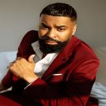 a photo of Ginuwine he is shown against a grey background wearing a red sport coat he is looking at the camera and has his left arm raised slightly to his chest