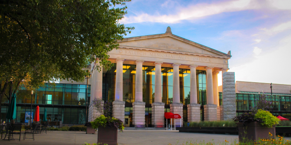 An exterior image of Raleigh Memorial Auditorium blue skies and clouds are shown in the top right corner and a tree with green leaves in the left corner